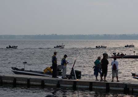 A line of boats idles out past the sailboats and into the horizon of Lake Champlain as fans look on. 