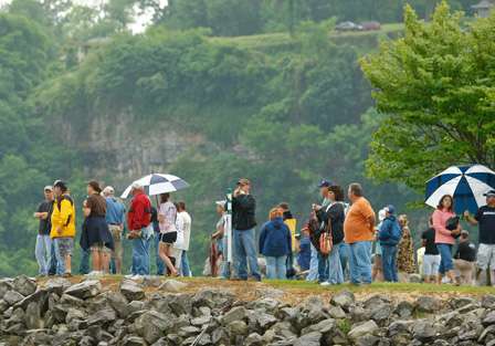 Fans stand along the shores of Pickwick Lake, huddled under umbrellas and wearing rain jackets while tornado sirens sound.